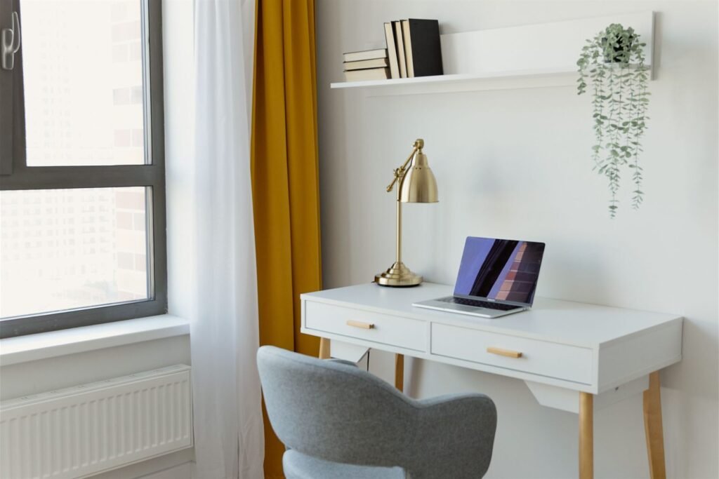 Bright minimal home office with a white desk, brass lamp, laptop, floating shelf and mustard curtain