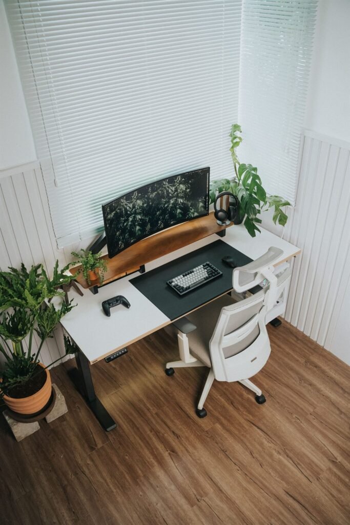 Corner home office with an electric sit-stand desk, curved monitor on a wooden shelf, white mesh chair and potted plants