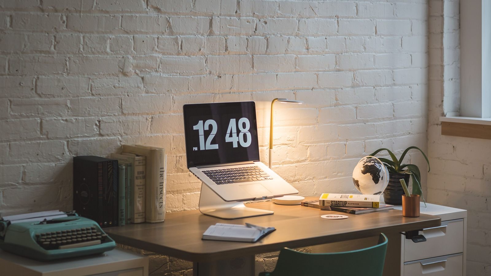 Desk against a white brick wall with a laptop on a stand, desk lamp, books, globe and vintage typewriter