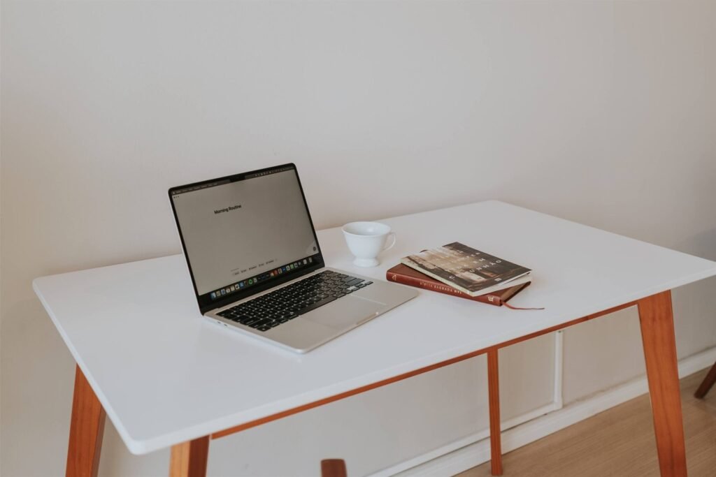 Minimal white desk with wooden legs, an open laptop, a coffee cup and two books