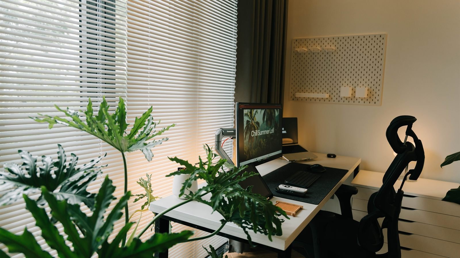 Modern home office with a monitor on an arm, pegboard wall, ergonomic chair and large plants by window blinds